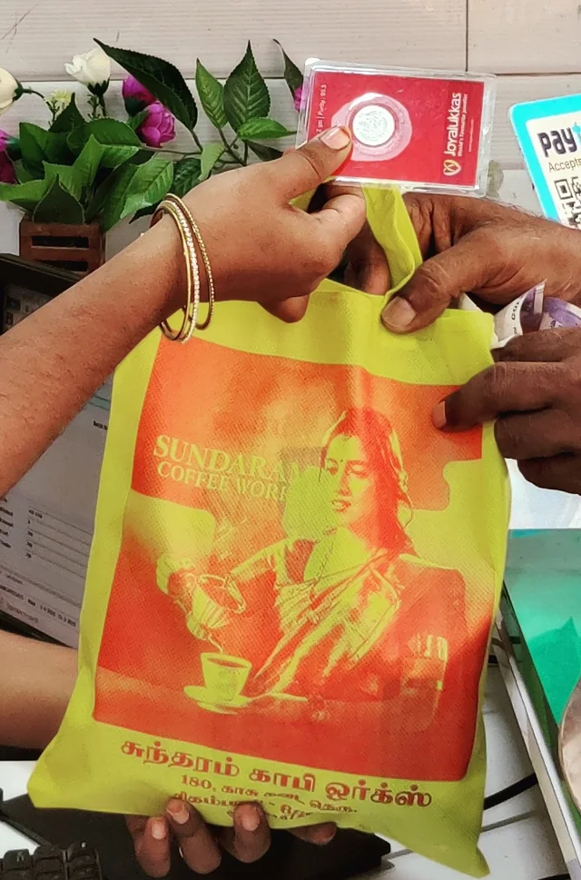 The Sundaram Coffee Works tote bag, printed with a woman pouring from a brass filter into a cup, above the shop name in English and Tamil