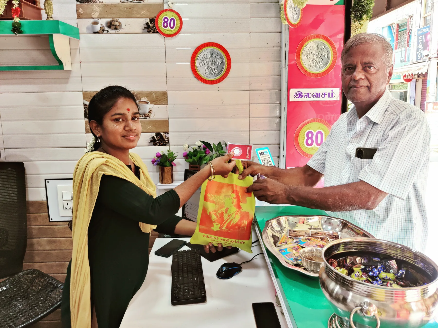 A Sundaram Coffee Works bag handed across the shop counter to a customer, Bazaar Street visible through the doorway